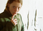 Young woman with finger on lower lip, blurred in foreground, peeling paint on wall in background