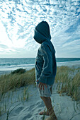 Boy standing on dune, wearing hooded sweatshirt, full length, side view