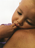 Child with fingers in mouth leaning against mother's bare shoulder