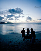 Fischgrillen auf kreolische Art mit Öltonnengrill am Strand am Abend, La Passe, im Hintergrund Nachbarinsel Praslin,  La Digue, La Digue and Inner Islands, Republik Seychellen, Indischer Ozean