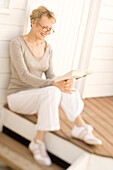 Smiling senior woman reading a book, sitting on wooden terrace