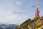 Junge Frau steht bei einem Gipfelkreuz in den Bergen, See, Tirol, Österreich