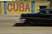 A blurred classic car passes a Viva Cuba sign, Havana, Cuba.
