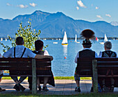 Couples wearing traditional clothes sitting on benches near harbor at lake Chiemsee, Prien, Chiemgau, Upper Bavaria, Germany