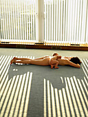 Mother and son lying naked on floor. Nude woman with baby boy lying on carpet floor. Sunlight is shining through venetian blinds.