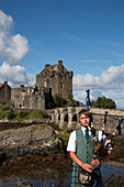Junger Mann im Schottenrock spielt Dudelsack vor Burg Eilean Donan Castle am Ufer von Loch Duich, nahe Dornie, Highland, Schottland, Großbritannien, Europa