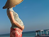 A pregnant woman stands on a beach, enjoying the morning sun