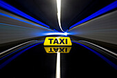 The yellow roof sign of a taxi. A cab driving through a tunnel. White and blue lights reflecting in the bodywork of the car.