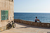 Rennradfahrer sitzt auf einer Mauer am Mittelmeer, Sant Elm, Mallorca, Balearische Inseln, Spanien