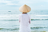 Frau mit vietnamesischer Kleidung und Strohhut blickt auf das Meer mit Fischerbooten, Strand von Mui Ne, Südvietnam, Vietnam, Asien