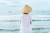 Woman wearing typical clothes and straw hat looking out to sea towards fishing boats, coast of Mui Ne, south Vietnam, Vietnam, Asia