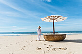Woman wearing typical clothes and straw hat looking out to sea towards fishing boats, coast of Mui Ne, south Vietnam, Vietnam, Asia