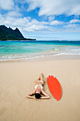 Hawaii, Kauai, Haena Beach Tunnels Beach, Woman laying on beach with surfboard.