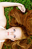 Junge Frau mit Sommersprossen und roten Haaren liegt im Gras und schaut auf.