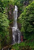 Hawaii, Maui, Kipahulu, Hana Coast, Woman stands with umbrella at Wailua Falls surrounded by foliage.