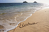 Hawaii, Oahu, Lanikai Beach aloha written in the sand with wave washing up onto shore, Mokulua islands and canoe in background