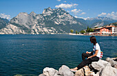 Junge Frau sitzt am Ufer des Gardasees, Torbole, Riva del Garda, Gardasee, Lago di Garda, Trient, Italien, Europa