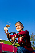 Junge Frau sitzt mit einem Glas Weißwein auf einem Traktor, Steiermark, Österreich