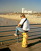 USA, California, Santa Monica, Los Angeles, a young man sits on a fire hydrant on the Santa Monica Pier with Santa Monica State Beach in the distance