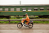 VIETNAM, Hanoi, Countryside, a man passes a train on his moped in the rain