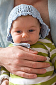 Baby with blue crocheted bonnet, being held by father in blue bath robe, Toronto, Ontario