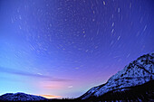Star trails above a mountain with northern lights on horizon, Yukon