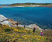 Hikers On The Coastal Path Around St Agnes With The Island Of Gugh And The Sand Bar In The Distance, Isles Of Scilly, Cornwall, Uk, Europe