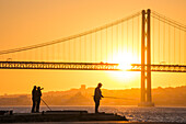 Silhouette of fishermen on Cacilhas waterfront with Ponte 25 de Abril bridge over Tagus river at sunset, Cacilhas, Almada (near Lisbon), Portugal