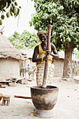 Woman carrying child on back grinding corn to flour, Magadala, Mali