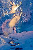 Snowman standing next to a stream with sunrays shining through hoar frosted trees in the background, Russian Jack Springs Park, Anchorage, Southcentral Alaska, Winter. Digitally enhanced.