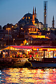 Fish restaurants being run from side of boats beside Golden Horn with Suleymaniye Mosque behind at dusk, Istanbul, Turkey