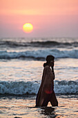 India, Karnataka, Indian girl walking in sea talking on mobile phone at sunset, Gokarna