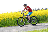Man riding an electric bicycle between blooming canola fields, Tanna, Thuringia, Germany