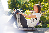 Frau sitzt auf einer Parkbank, Beine über der Lehne, München, Bayern, Deutschland