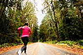 A female jogging down a road next to tall trees covered in moss Port Angeles, Washington, USA