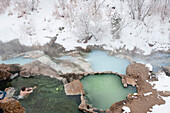 A young man sits in a natural hot springs on a snowy day in Springville, Utah.  The pools are three different shades of green Springville, Utah, USA