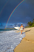 An amazing rainbow over Rocky Point, as a surfer girl walks along the beach, on the north shore of Oahu, Hawaii north shore, Oahu, Hawaii, USA