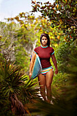 young woman walking through thick vegetation with surfboard.  going to beach to surf Grayton Beach, Florida, United States