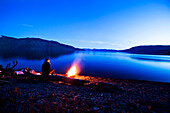 A woman sits next to a beautiful campfire at sunset in Idaho Sandpoint, Idaho, USA