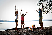 Three young adults smiling and dancing around a camp fire on a camping and kayaking trip on a lake in Idaho Sandpoint, Idaho, USA