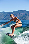 A athletic young woman smiles while wakesurfing behind a wakeboard boat on a sunny day in Idaho., Sandpoint, Idaho, USA