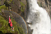 A woman standing with arms raised under a rainbow and next to waterfall, Rio Grande National Forest, Creede, Colorado., Creede, Colorado, usa