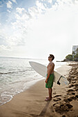 Japanese man holding surfboard on beach, Honolulu, HI