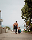 Caucasian father and son in wetsuits walking toward the ocean, Manhattan Beach, California, USA