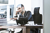 Businessman sitting at desk with computer and paperwork, using smartphone