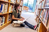 An 18 year old girl sitting on the floor in a library with a laptop computer and books stacked next to her.