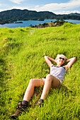 'A Young Woman Lays On The Grass With A View Of The Coastline; Urupukapuka Island, New Zealand'