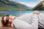 'A Girl Lies Down On A Picnic Table And Enjoys The Sun With A Sailboat In Lake Rotoiti In The Background, Tasman Region; New Zealand'
