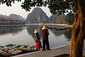 Two women with typical vietnamese conic hats talks on the bank of the river at sunset in Tam Coc with carsic mountains in the background, Ninh Binh, Vietnam