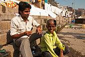 Barber cuts a green shirt dressed boy's hair on the banks of the Ganges river at sunrise in Varanasi, India